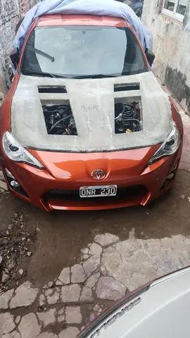 El capó de fibra en crudo montado sobre el auto naranja, con el motor visible por las aberturas.