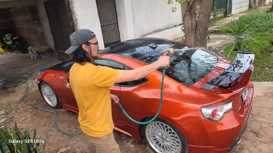 El Toyota 86 naranja recién lavado en el patio, ya con el capó negro, el alerón trasero y el techo oscuro, mientras lo enjuagan con una manguera.