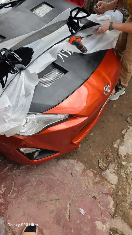 El capó negro ya montado sobre el auto naranja, todavía con plástico de protección, mientras se atornilla con un destornillador eléctrico.