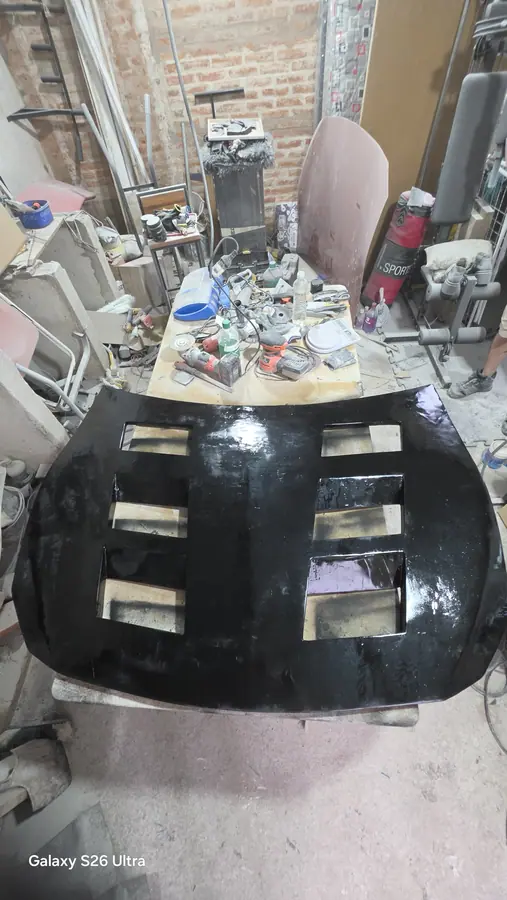El capó de fibra de vidrio terminado en negro brillante, visto de frente sobre los caballetes del taller.