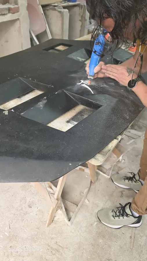 Pedro grabando a mano con una herramienta rotativa sobre el capó negro terminado, con el polvo blanco del grabado saltando.