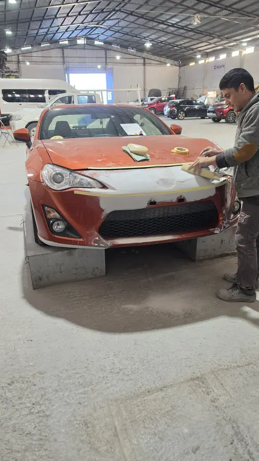 Un operario lija el frente enmascarado del auto en el taller, con el paragolpes en imprimación gris.